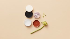 Artistic flat lay of makeup containers, powder balls, and flower buds on a soft yellow surface.