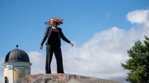 Young woman posing confidently on a rock in a Helsinki park, with a clear blue sky and European architecture in the background.
